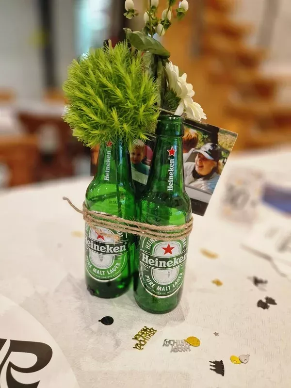 Zwei grüne Heineken-Bierflaschen, mit Juteschnur zusammengebunden und als Vase für ein Blumenarrangement mit grüner Deko und weißen Blüten genutzt, stehen als Tischdekoration auf einem festlich gedeckten Tisch